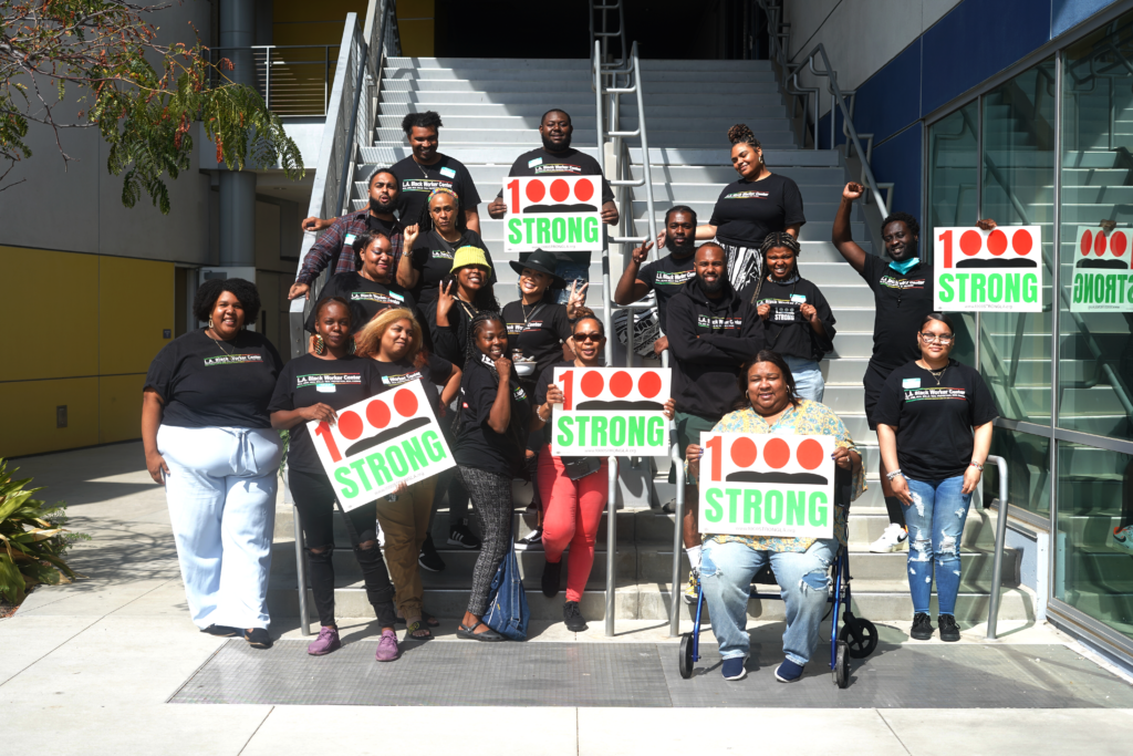 Group of 19 Black folks posing in front of a staircase, smiling, and holding signs that say "1000 Strong"