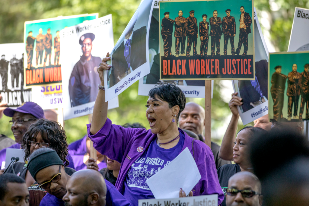 A Black worker with short hair wearing purple and standing in a crowd holds up a sign
