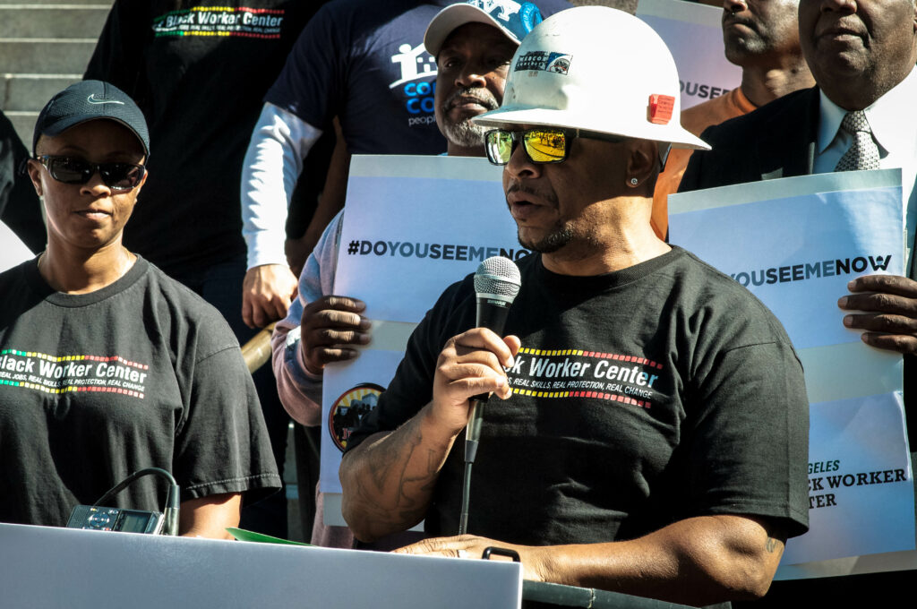 A Black worker wearing a hard hat and sunglasses speaks into a microphone while a group of Black workers stand behind them holding signs.