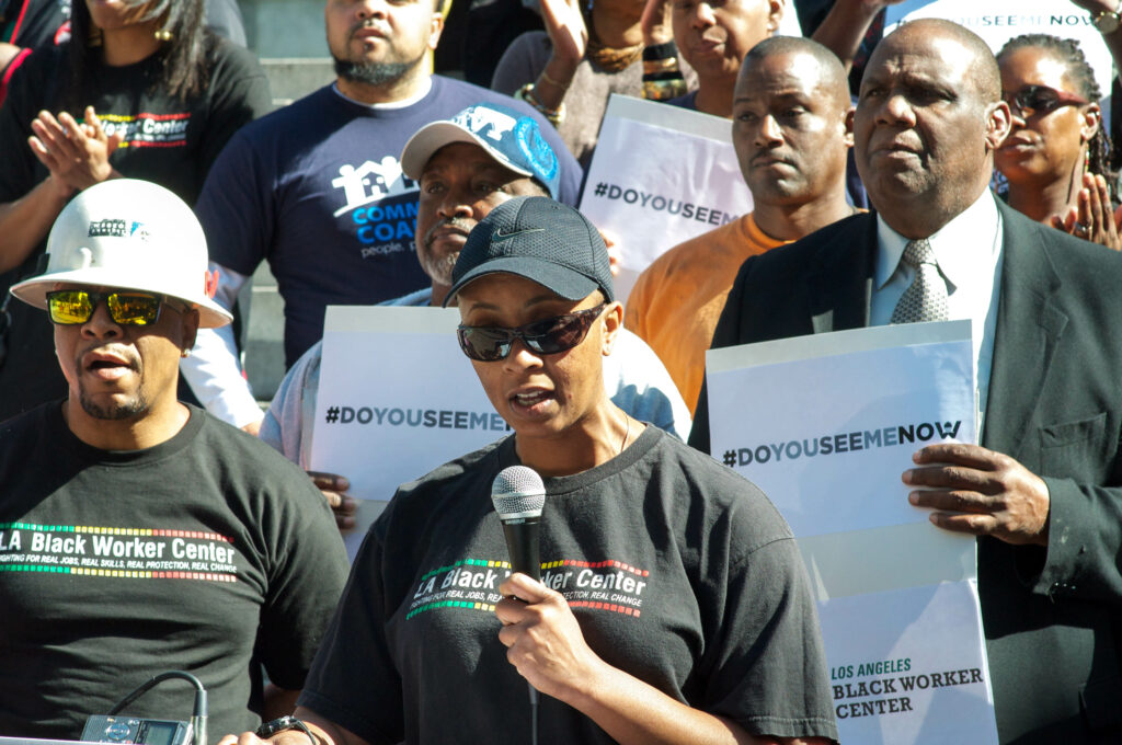 A Black worker wearing a hat and sunglasses speaks into a microphone while a group of Black workers stand behind them holding signs.