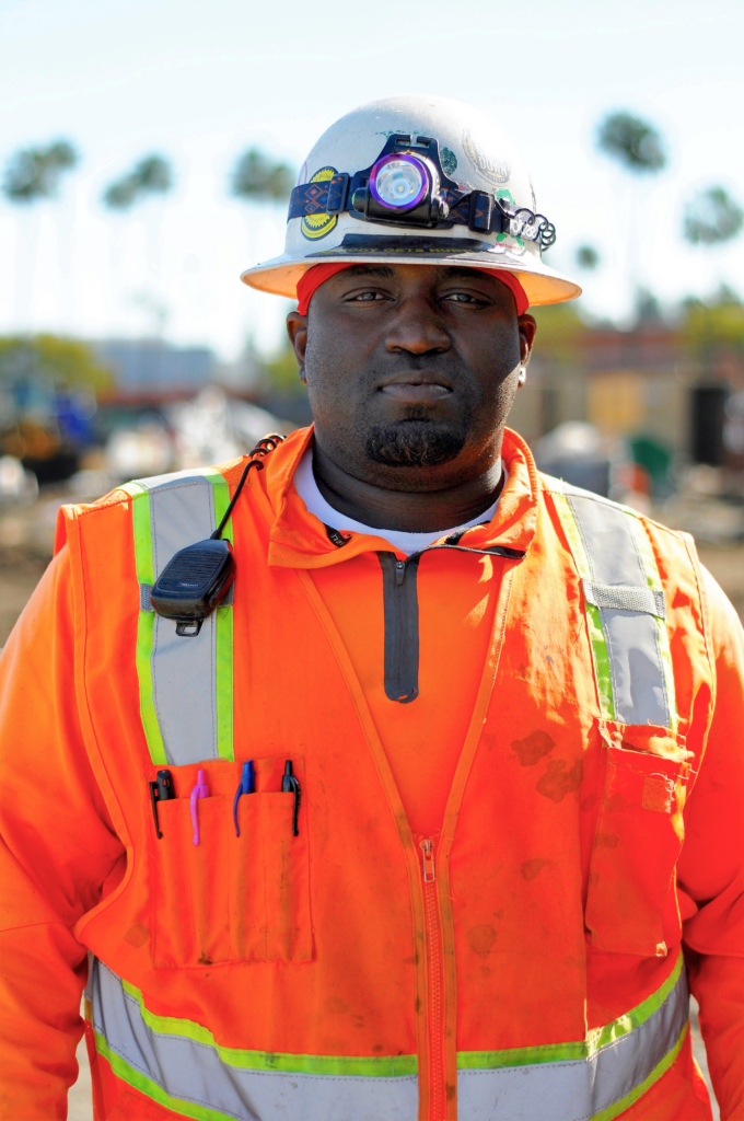 A Black worker wearing an orange hard hat, vest, and long sleeve shirt looks straight into the camera with a soft smile