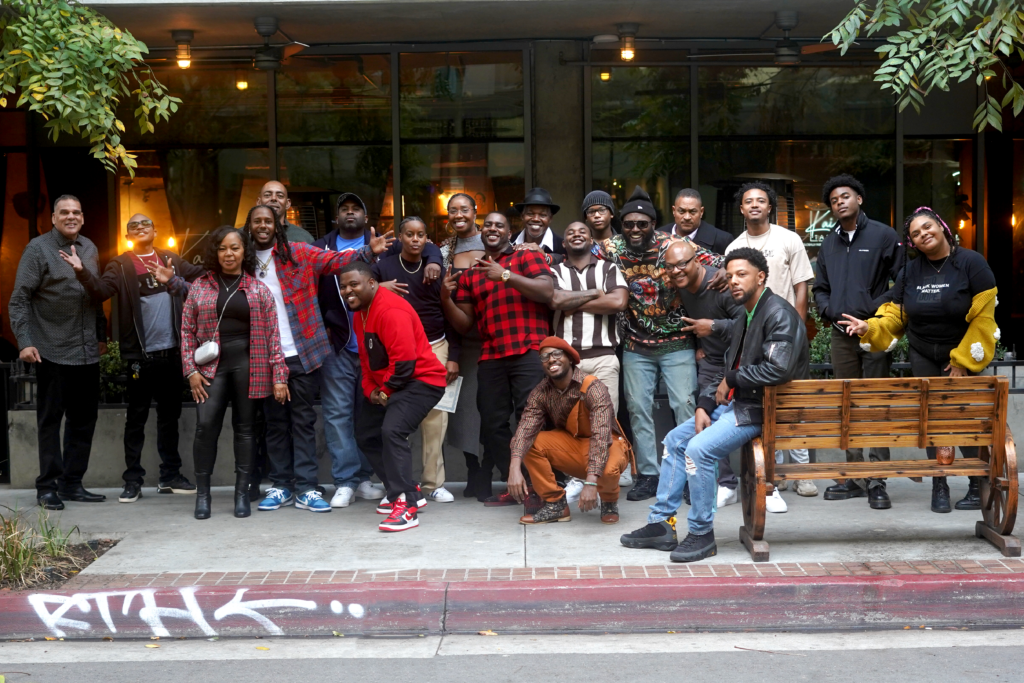 A group of over 20 Black folks posing in front of a camera on the sidewalk and smiling