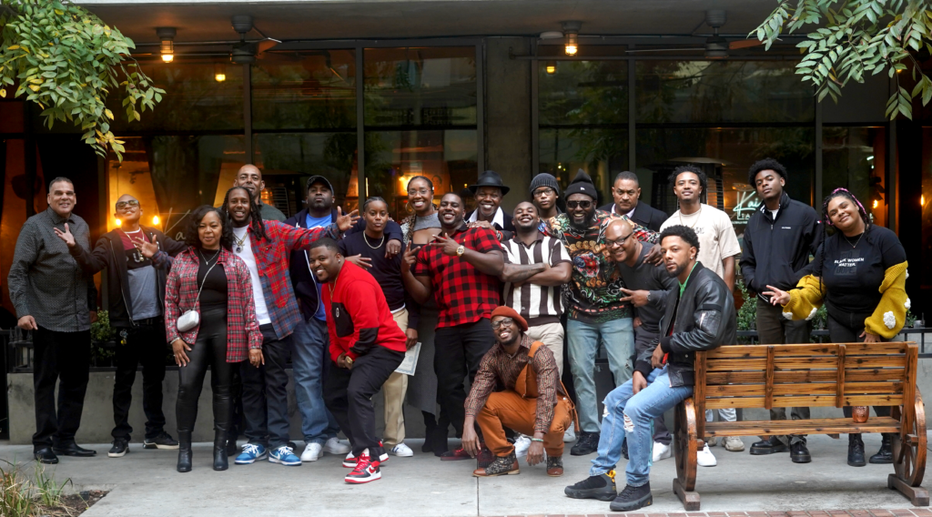 A group of over 20 Black folks posing in front of a camera on the sidewalk and smiling