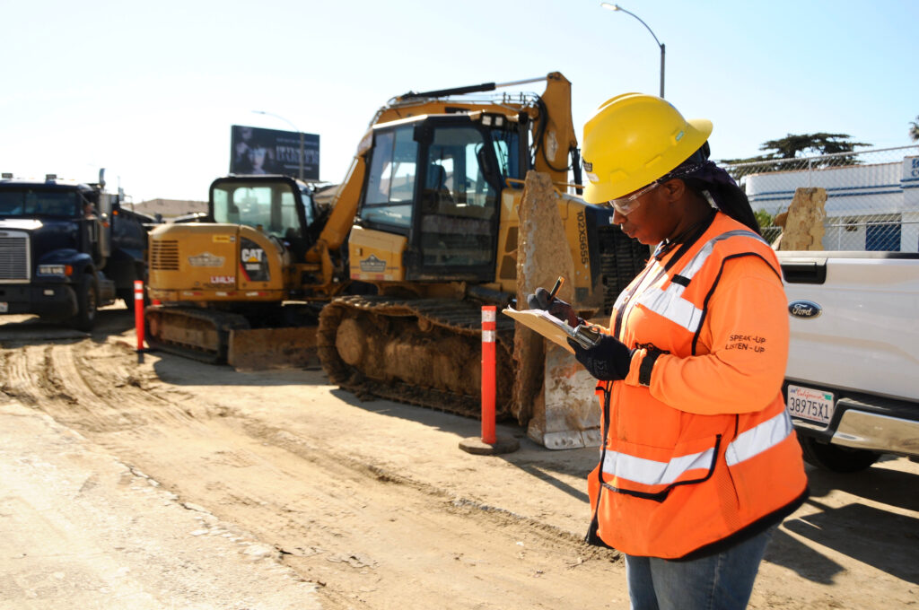 A Black worker with long hair wearing a hard hat and orange vest looks down at a clipboard while standing in front of a yellow tractor.