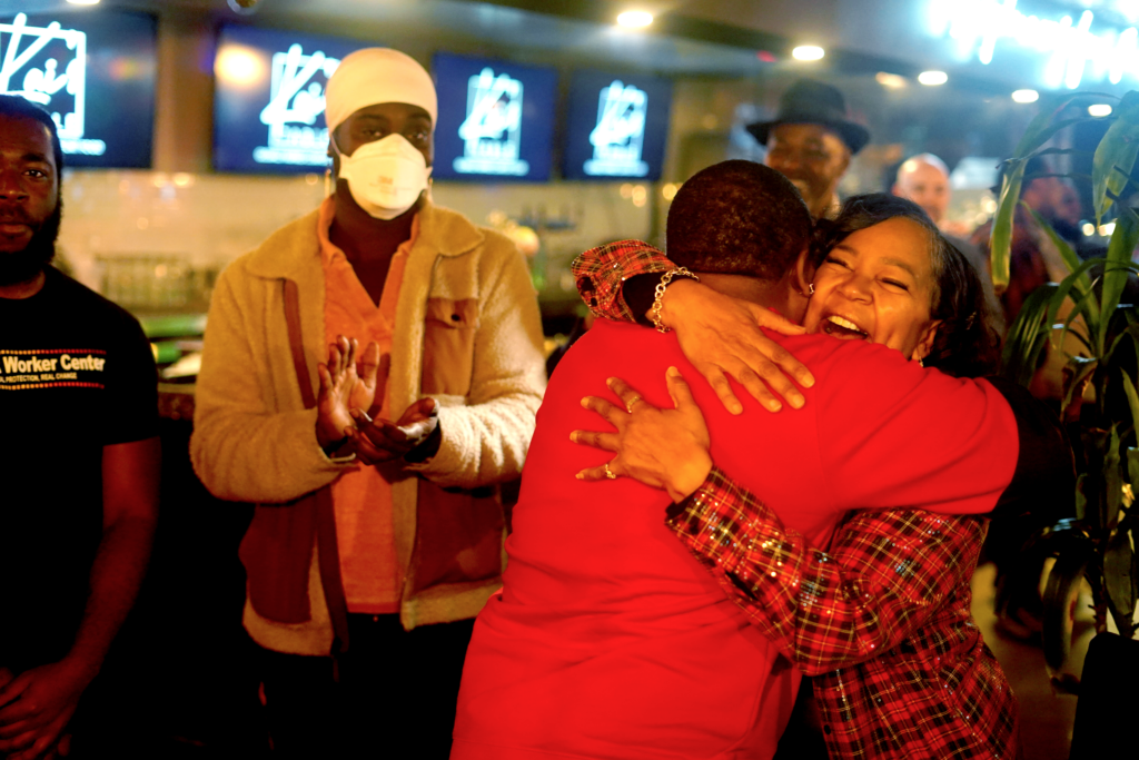 A Black woman happily hugs another person who is wearing red. A Black worker wearing a mask stands to the side of them clapping their hands.