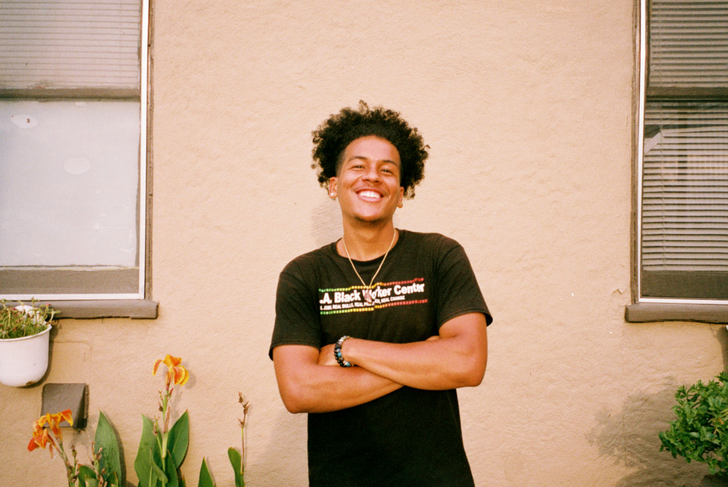 A young Black worker with short hair and their arms crossed smiles in front of the camera