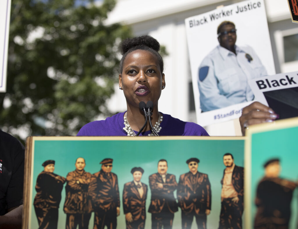 A Black person with their hair tied up speaks behind a microphone. There are posters around them including one that says "Black Worker Justice"