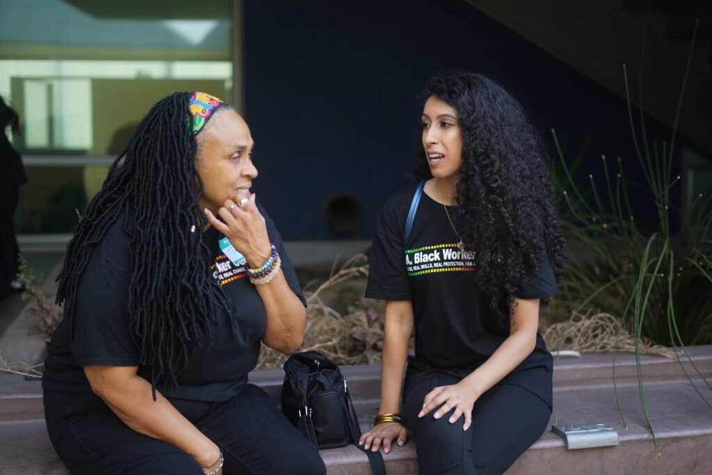 Two Black workers with long hair are sitting and talking to each other