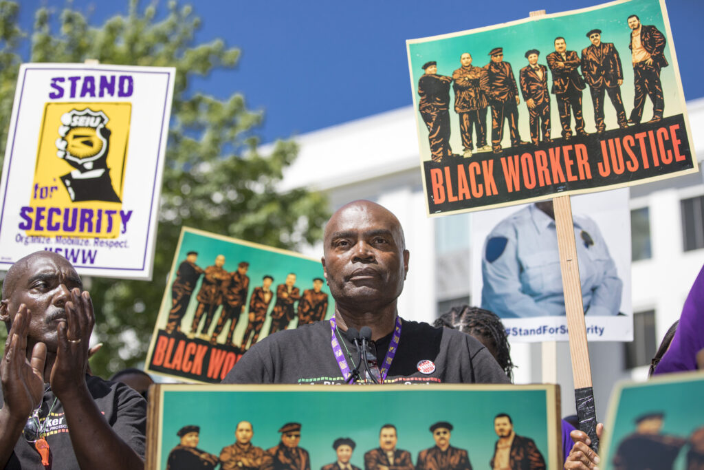 A Black worker standing behind a microphone looks sternly into the crowd. There are signs around them featuring a group of workers and the words "Black worker--"